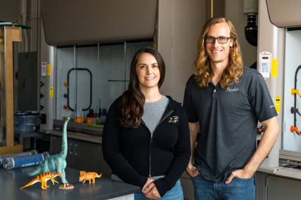 Photo of Alexandria Johnson and Brandon Johnson in a laboratory. They're casually dressed and there are some model dinosaurs sitting on a lab bench next to them. There's a fume hood in the background.