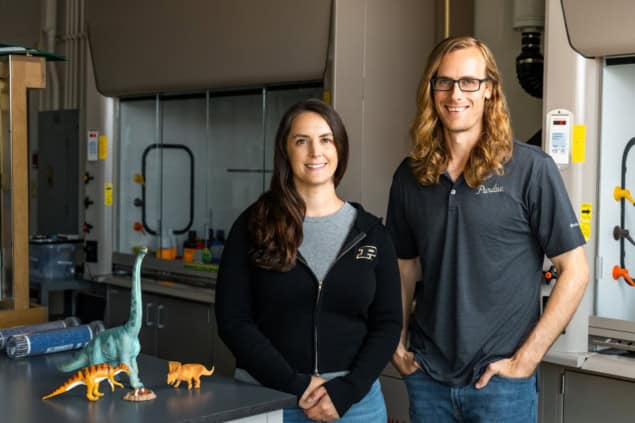 Photo of Alexandria Johnson and Brandon Johnson in a laboratory. They're casually dressed and there are some model dinosaurs sitting on a lab bench next to them. There's a fume hood in the background.