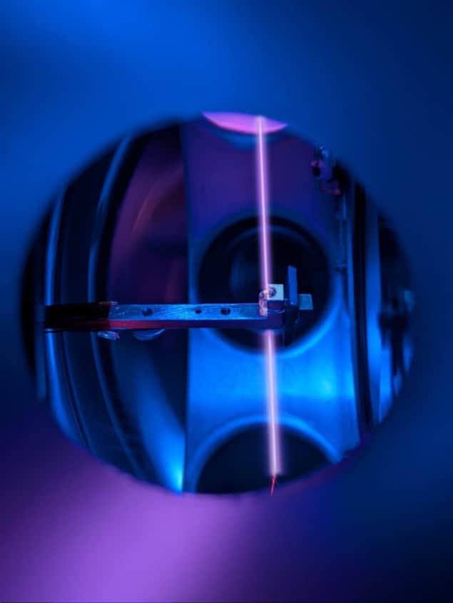A purplish laser beam passing through a small vial, viewed through a port in a vacuum system