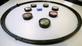 Photo showing eight bristlebots confined by a black circular ring. Seven of the bristlebots are surrounded by plastic rings and covered in semi-transparent foil. The eighth bristlebot is unmodified and is bright blue.