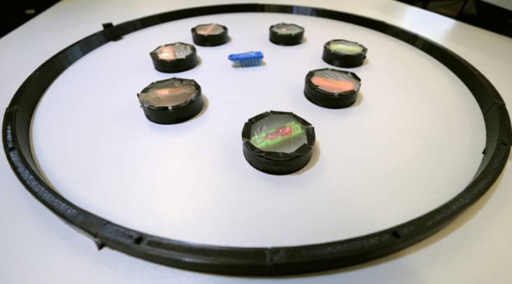 Photo showing eight bristlebots confined by a black circular ring. Seven of the bristlebots are surrounded by plastic rings and covered in semi-transparent foil. The eighth bristlebot is unmodified and is bright blue.