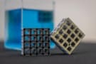 Two structures made of a lattice of cubic voids. One set of voids is filled, the other isn't. A larger cube filled with blue liquid or gel is in the background