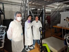 Photo of three researchers in white lab coats standing near blocks of shielding. The first researcher wears a face mask and a hair net, the second one wears booties and is flashing a peace sign with a purple nitrile gloved hand, and the third has no additional protective gear