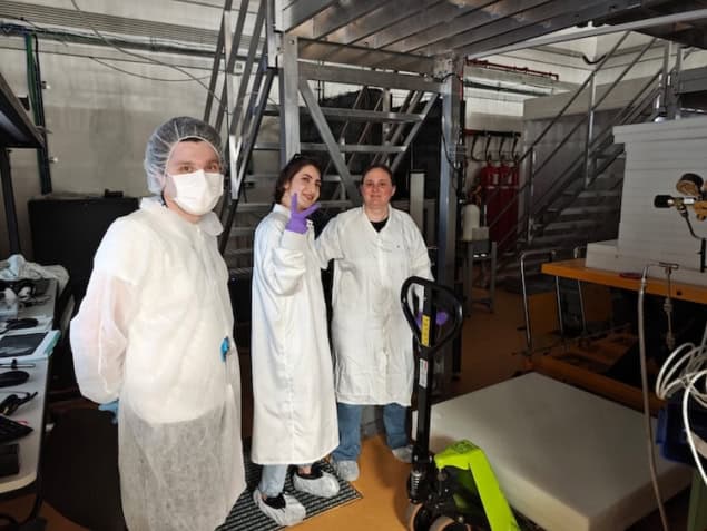 Photo of three researchers in white lab coats standing near blocks of shielding. The first researcher wears a face mask and a hair net, the second one wears booties and is flashing a peace sign with a purple nitrile gloved hand, and the third has no additional protective gear